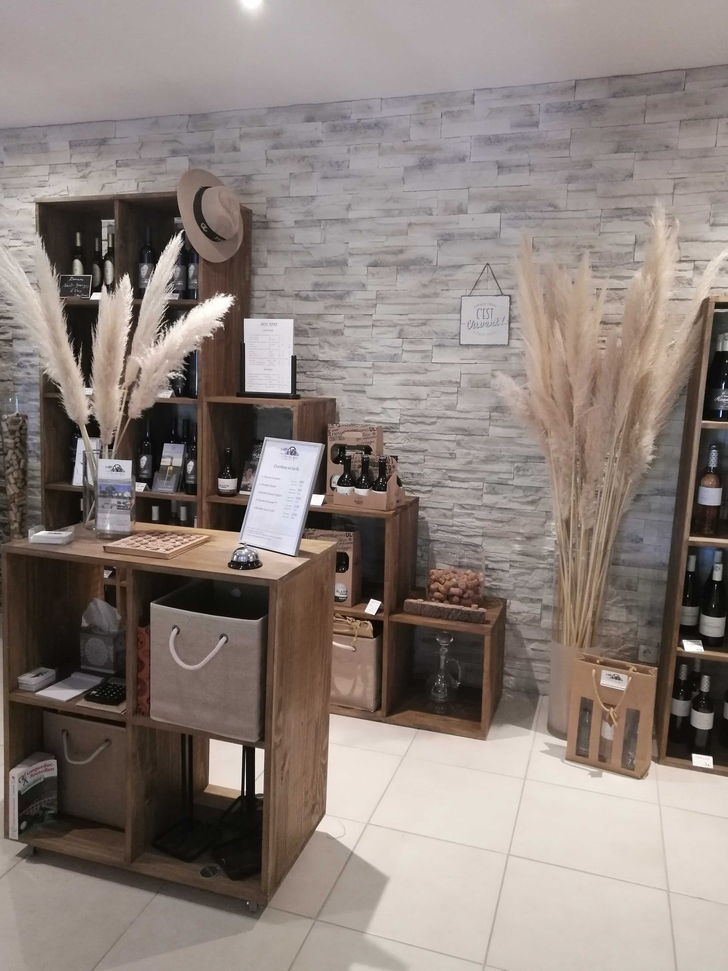 Intérieur d'une boutique avec présentoirs en bois, bouteilles de vin, panneaux de dégustation et grands bouquets de roseaux séchés devant un mur en pierre.